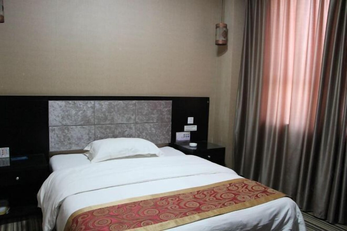 A hotel room with a bed, a nightstand, a wall clock, and a window with curtains.