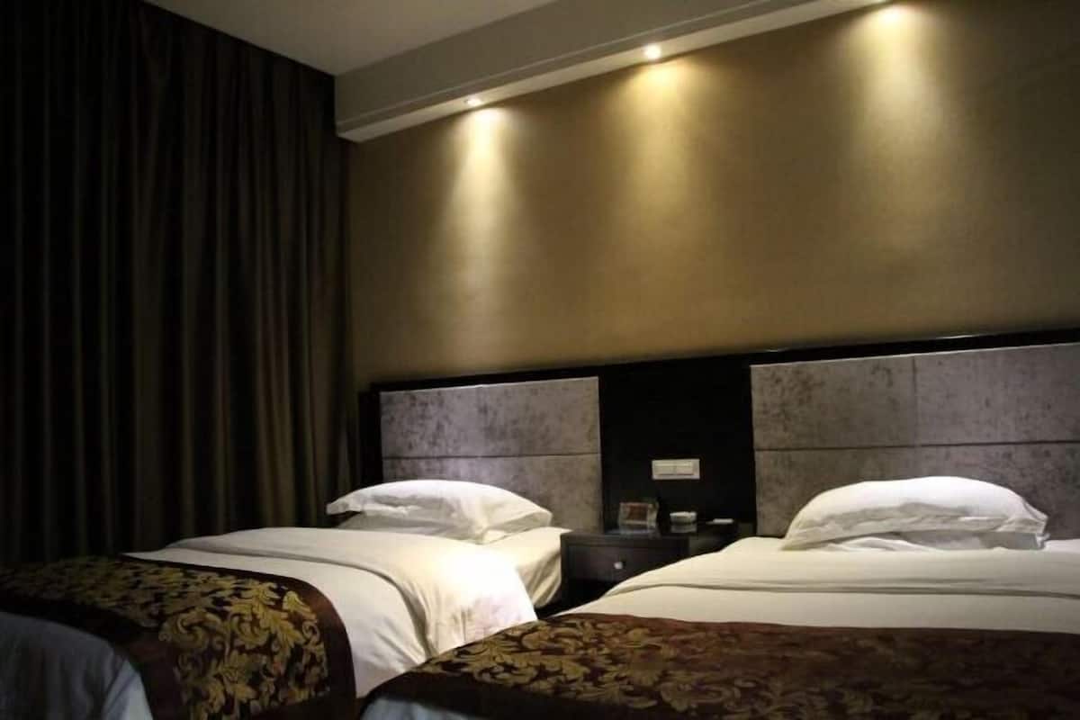 A hotel room with two beds, dark curtains, a headboard with a light fixture, and a nightstand with a lamp.