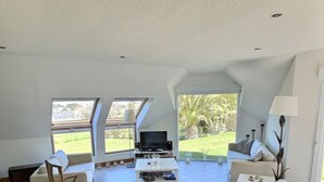 A spacious living room with a high ceiling, large windows, and a television.