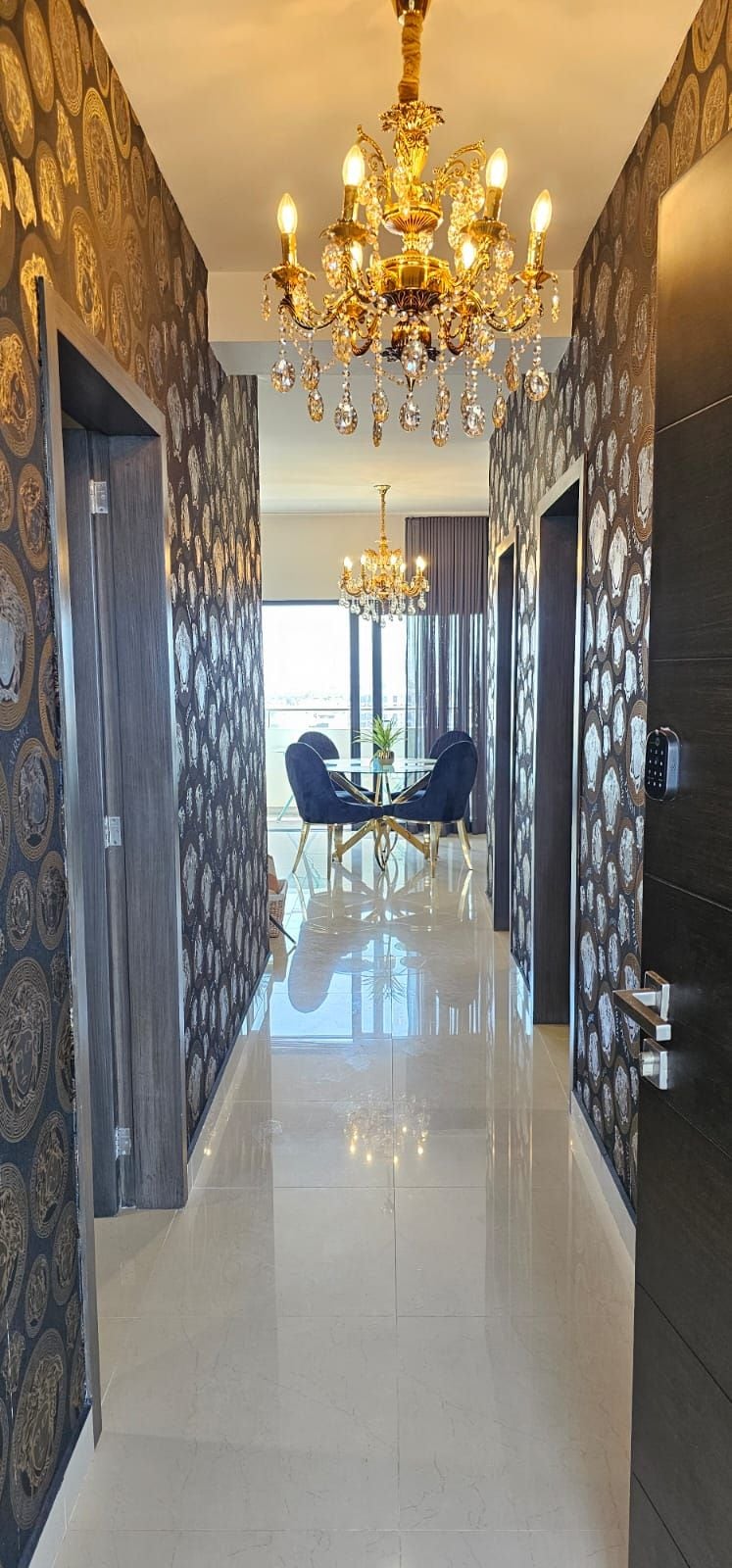 A hallway with a chandelier, patterned wallpaper, and a dining area with chairs.