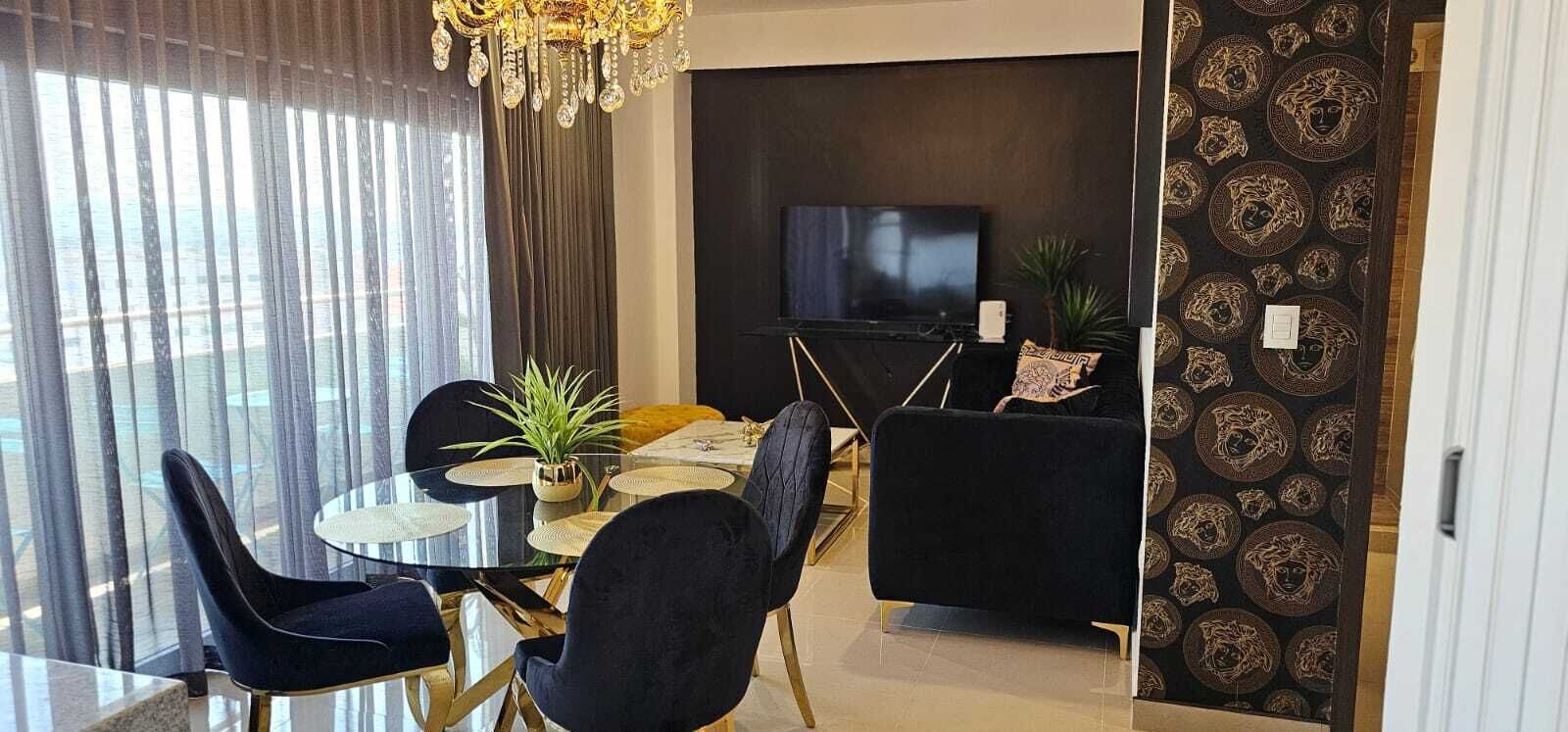 A dining area with a round table, black chairs, a chandelier, and a TV in the background.