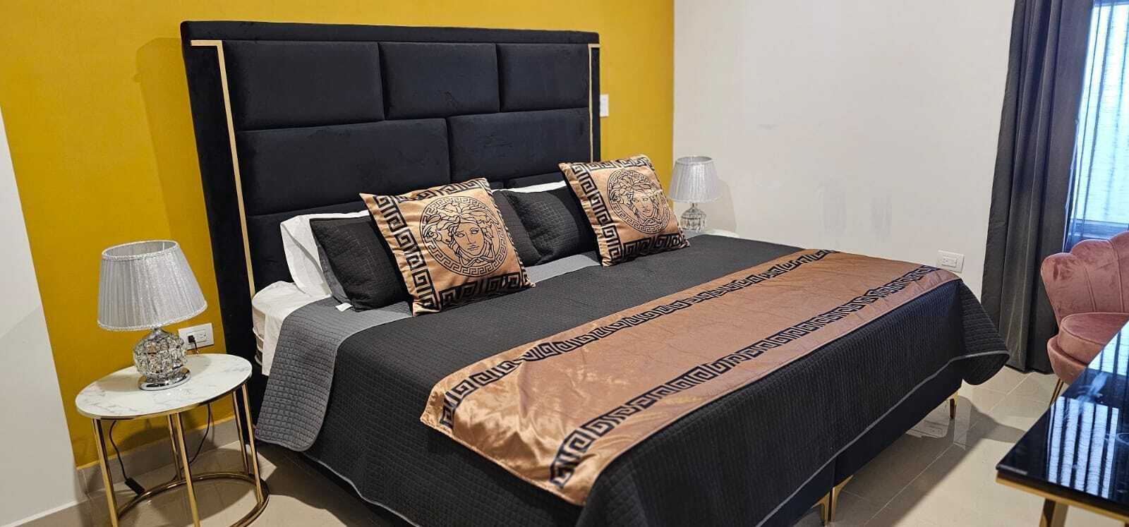 A hotel room with a black upholstered headboard, a bed with patterned pillows, a grey blanket, and a small round table with a lamp.