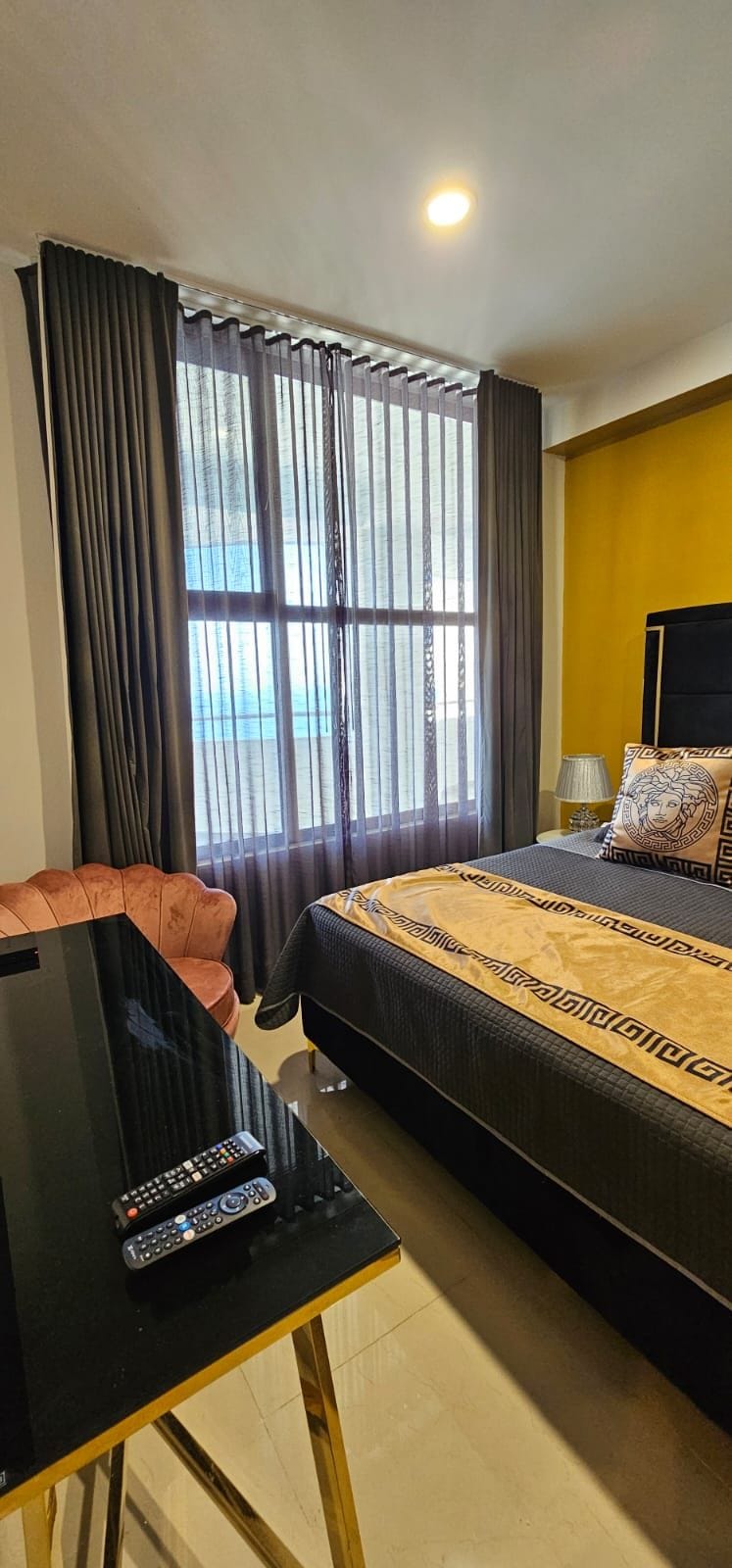 Una habitación de hotel con cama, escritorio con control remoto, silla y ventana con cortinas.
