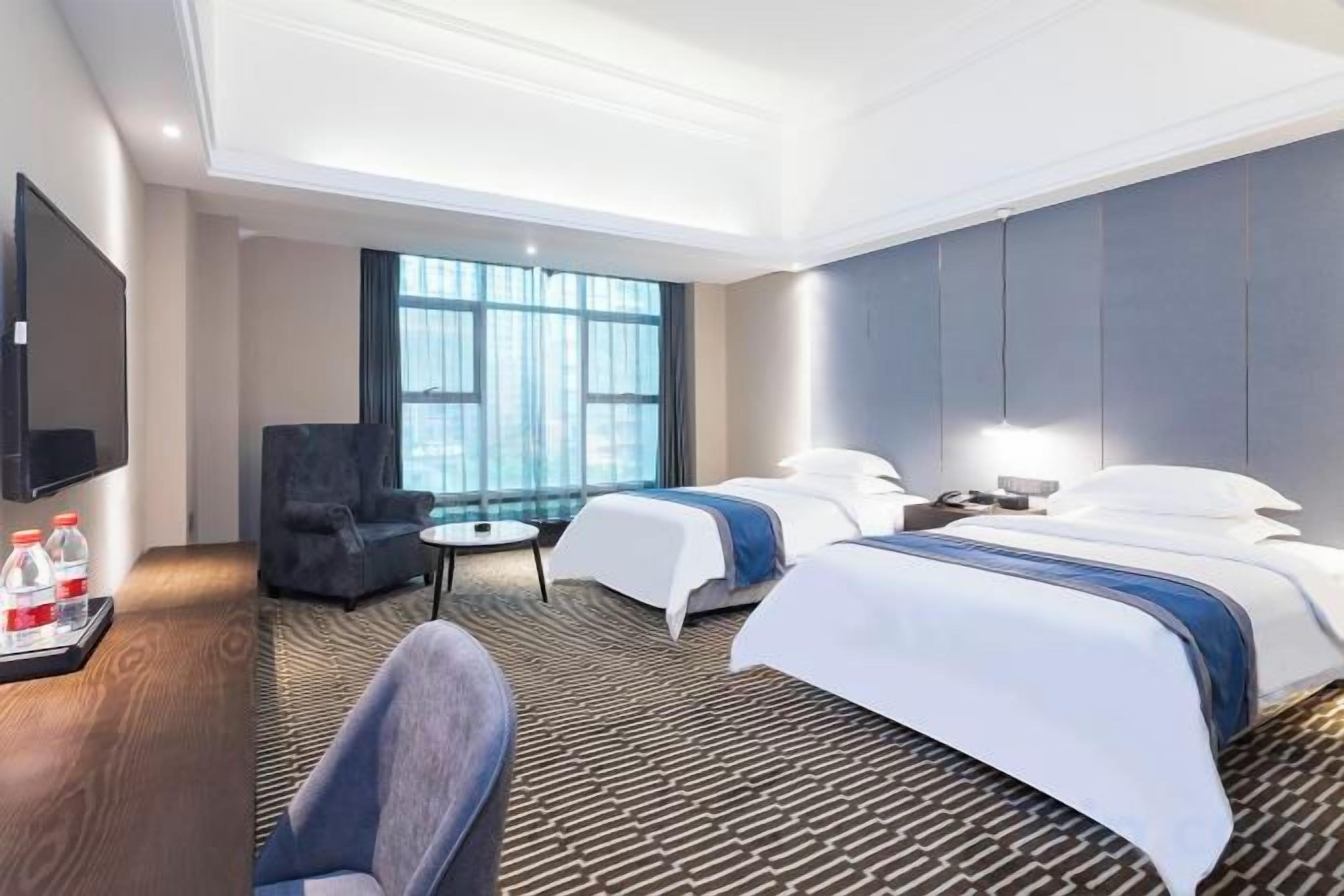 Una habitación de hotel con dos camas, un escritorio con silla, televisor y ventana con cortinas.