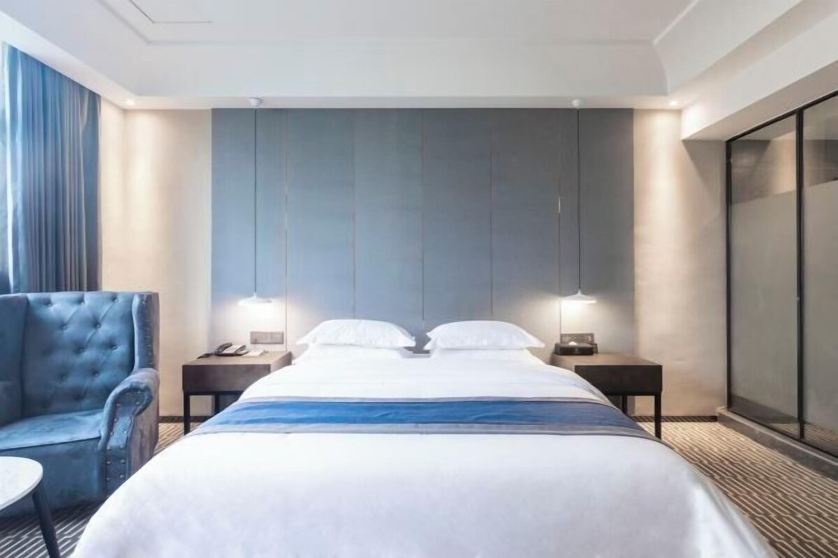 A hotel room with a large bed, two bedside tables with lamps, a blue armchair, and a large window with curtains.