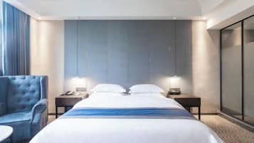 A hotel room with a large bed, two bedside tables with lamps, a blue armchair, and a large window with curtains.