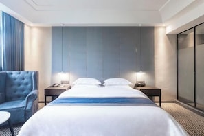 A hotel room with a large bed, two bedside tables with lamps, a blue armchair, and a large window with curtains.