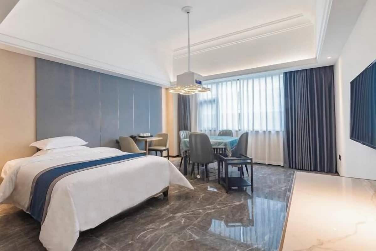 A hotel room with a bed, a desk, and a chair. There is a window with curtains, a ceiling light, and a TV on the wall.