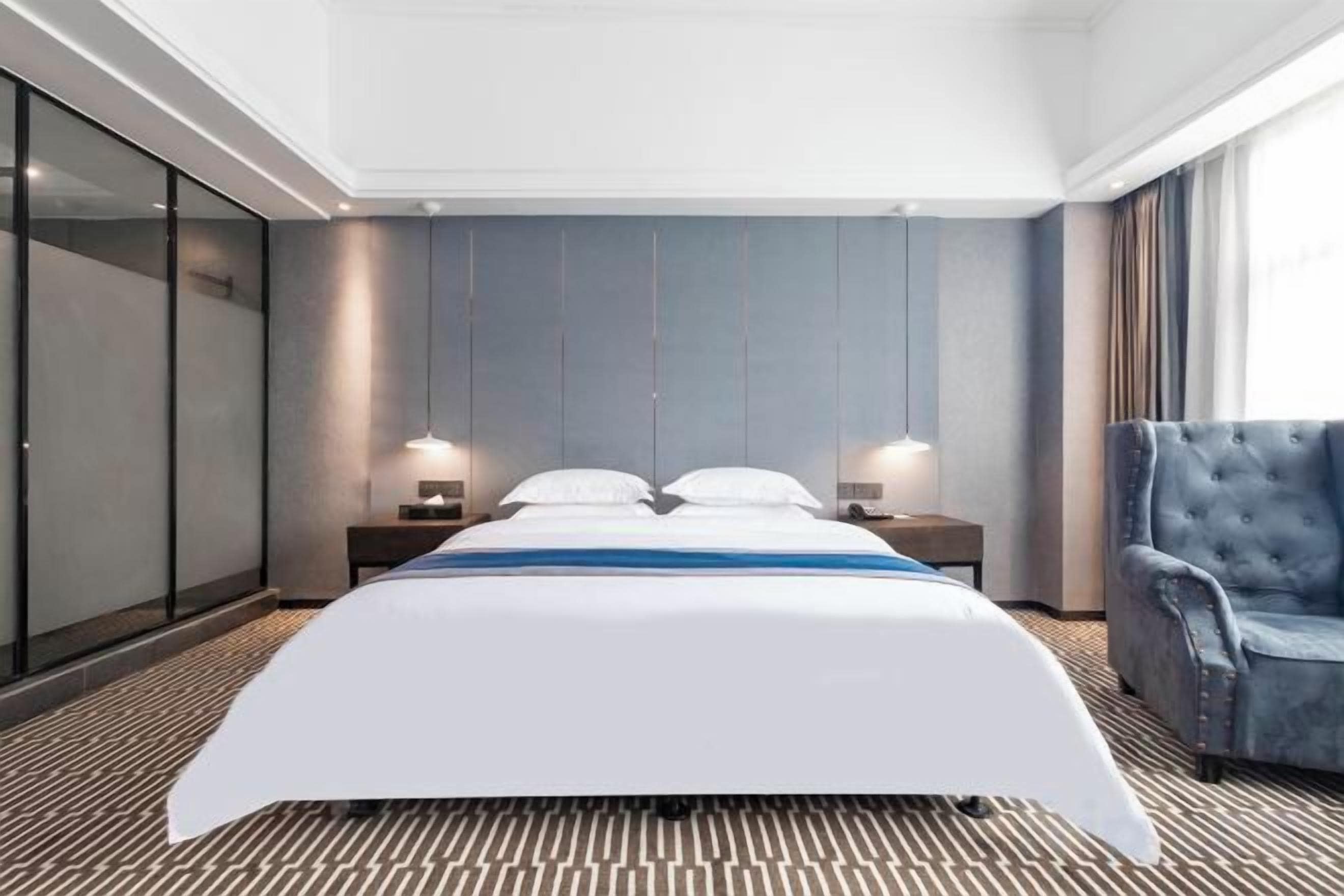 A hotel room with a large bed, two bedside tables, a blue armchair, and a large window.