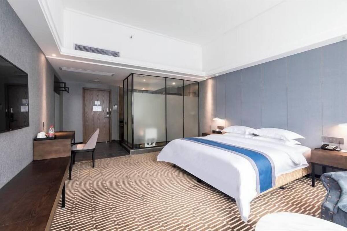 A modern hotel room with a large bed, a desk, a chair, and a glass-enclosed bathroom.