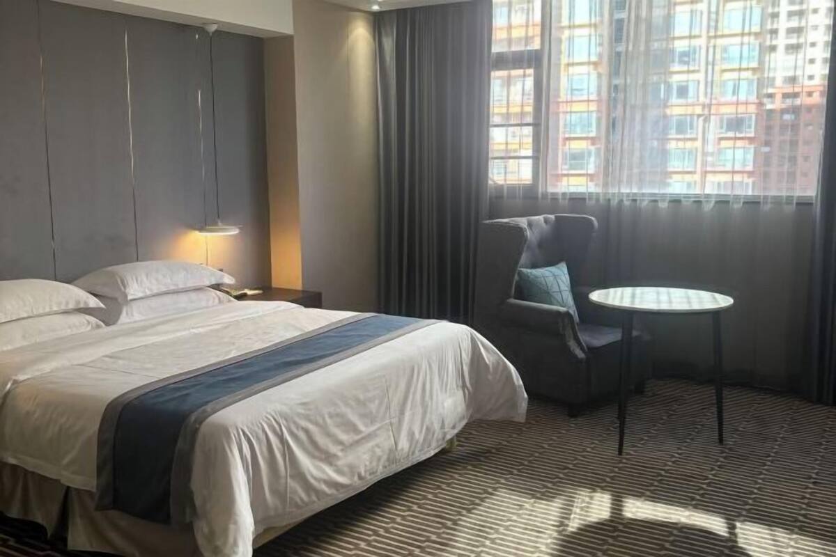 A hotel room with a bed, a chair, a small table, and a window with curtains.