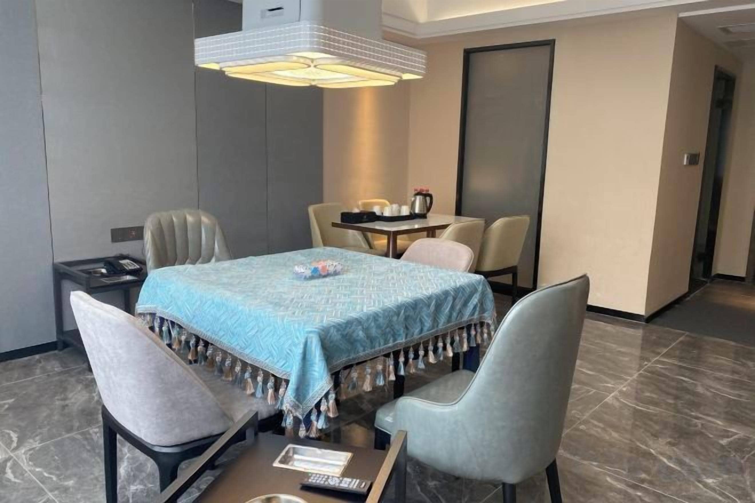 A hotel room with a dining table, two chairs, a small table with a kettle, and a bed with a headboard.