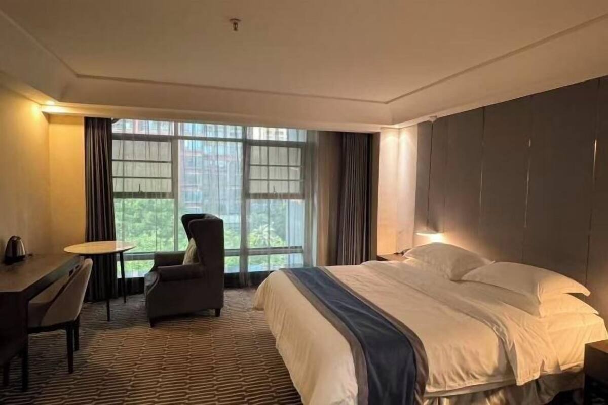 A hotel room with a bed, a desk, a chair, and large windows.