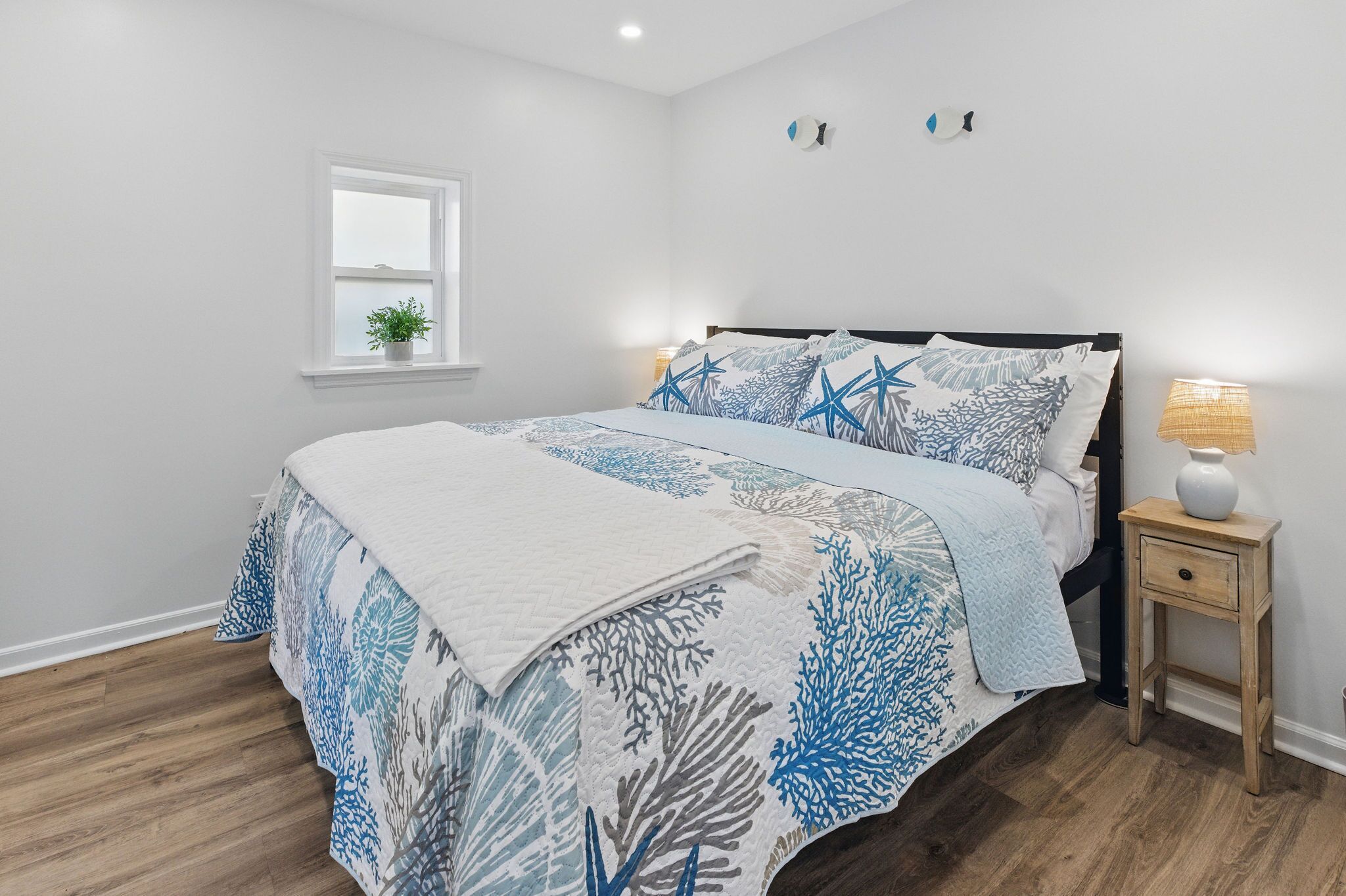 A bedroom with a bed, a nightstand, a lamp, a window, and a wall-mounted light fixture.