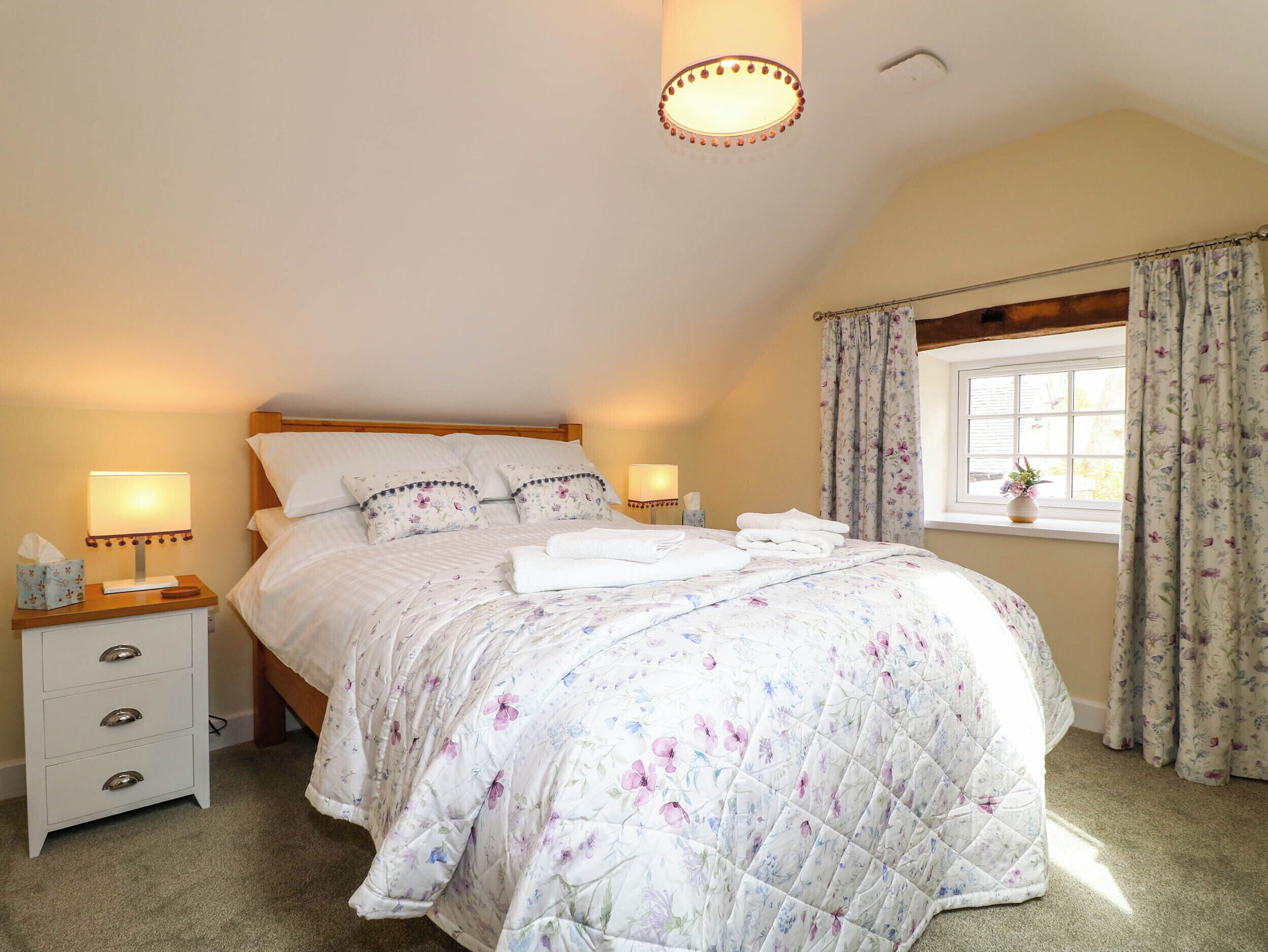 A neatly made bed with floral bedding, two bedside lamps, a nightstand with drawers, and a window with curtains.