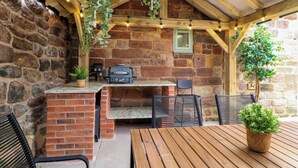 Cocina al aire libre con paredes de piedra, encimera de ladrillo, plantas colgantes y una parrilla.