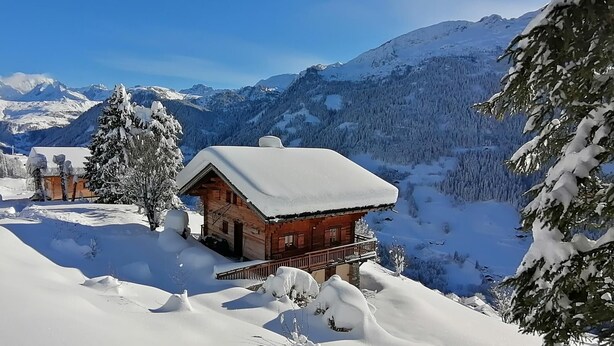 A wooden cabin with a snow-covered roof, surrounded by snow-covered trees and mountains in the background.