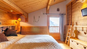 A cozy wooden room with a bed, a dresser with heart-shaped cutouts, a window with a view of mountains, and a wooden ceiling.