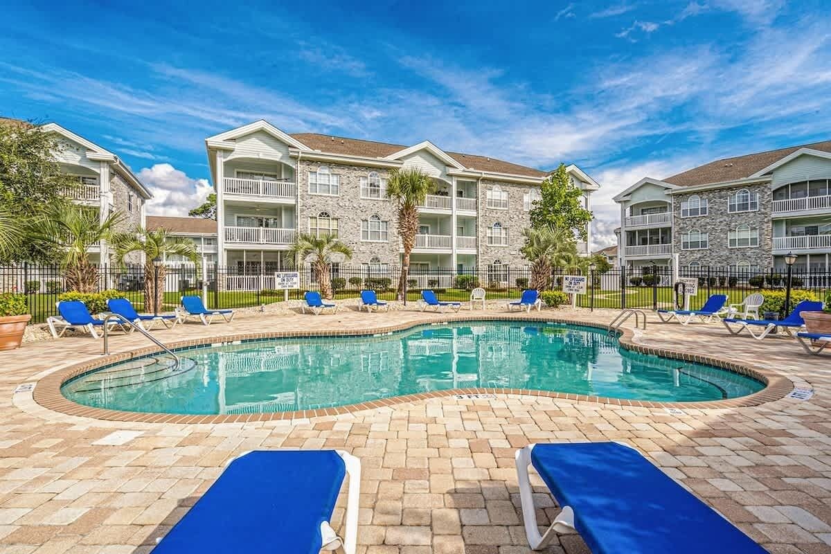 A pool area with lounge chairs, a swimming pool, and multi-story buildings with balconies.