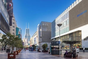 Una amplia calle urbana con edificios modernos, cielo despejado y algunos peatones.