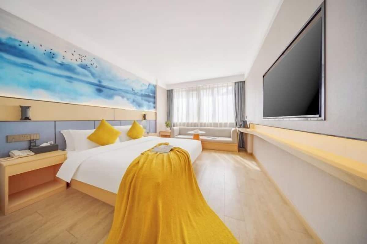 A hotel room with a bed, bedside table, lamp, artwork, window, and a TV.