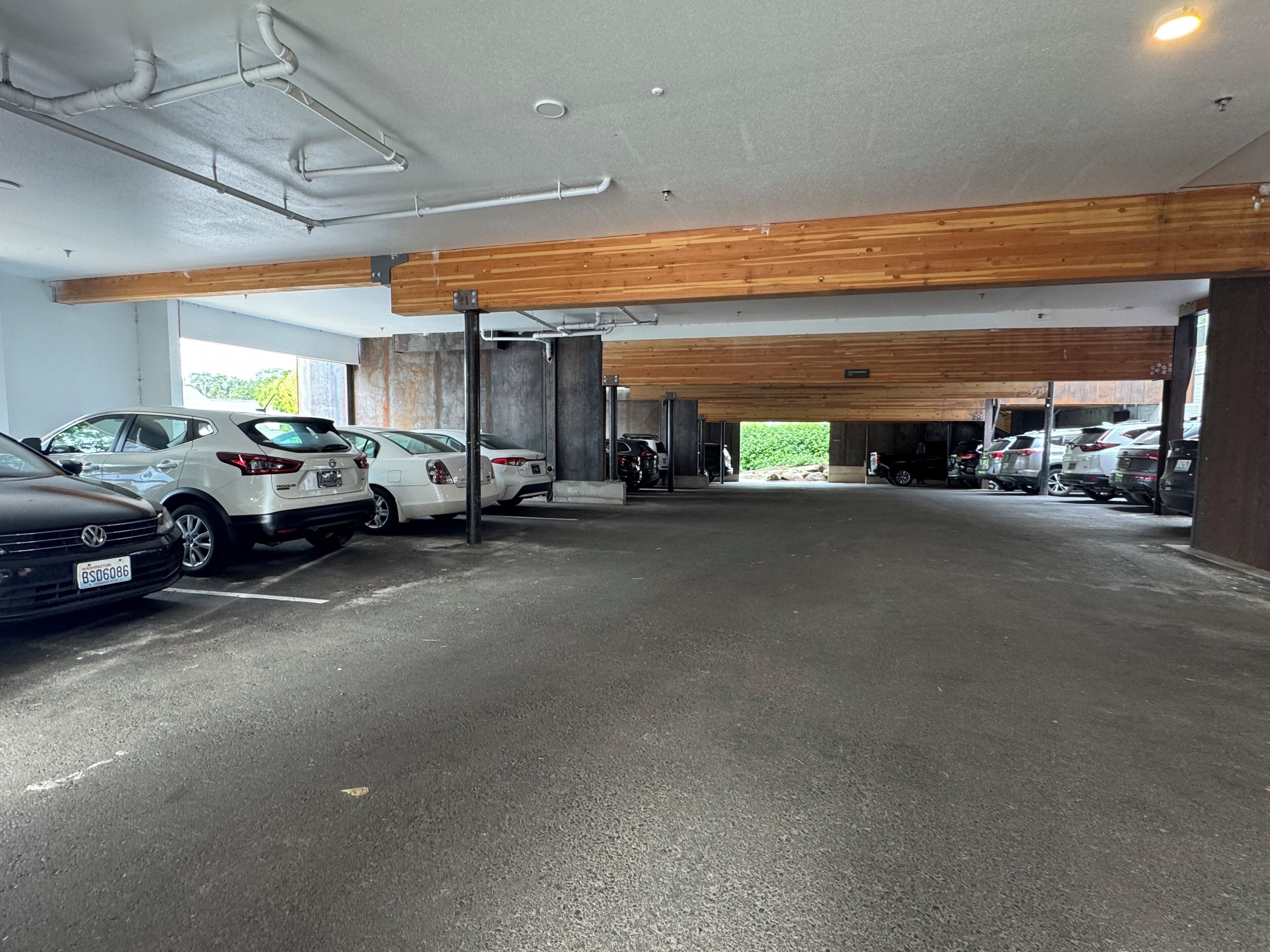 A parking garage with multiple cars parked, concrete floors, and wooden beams on the ceiling.
