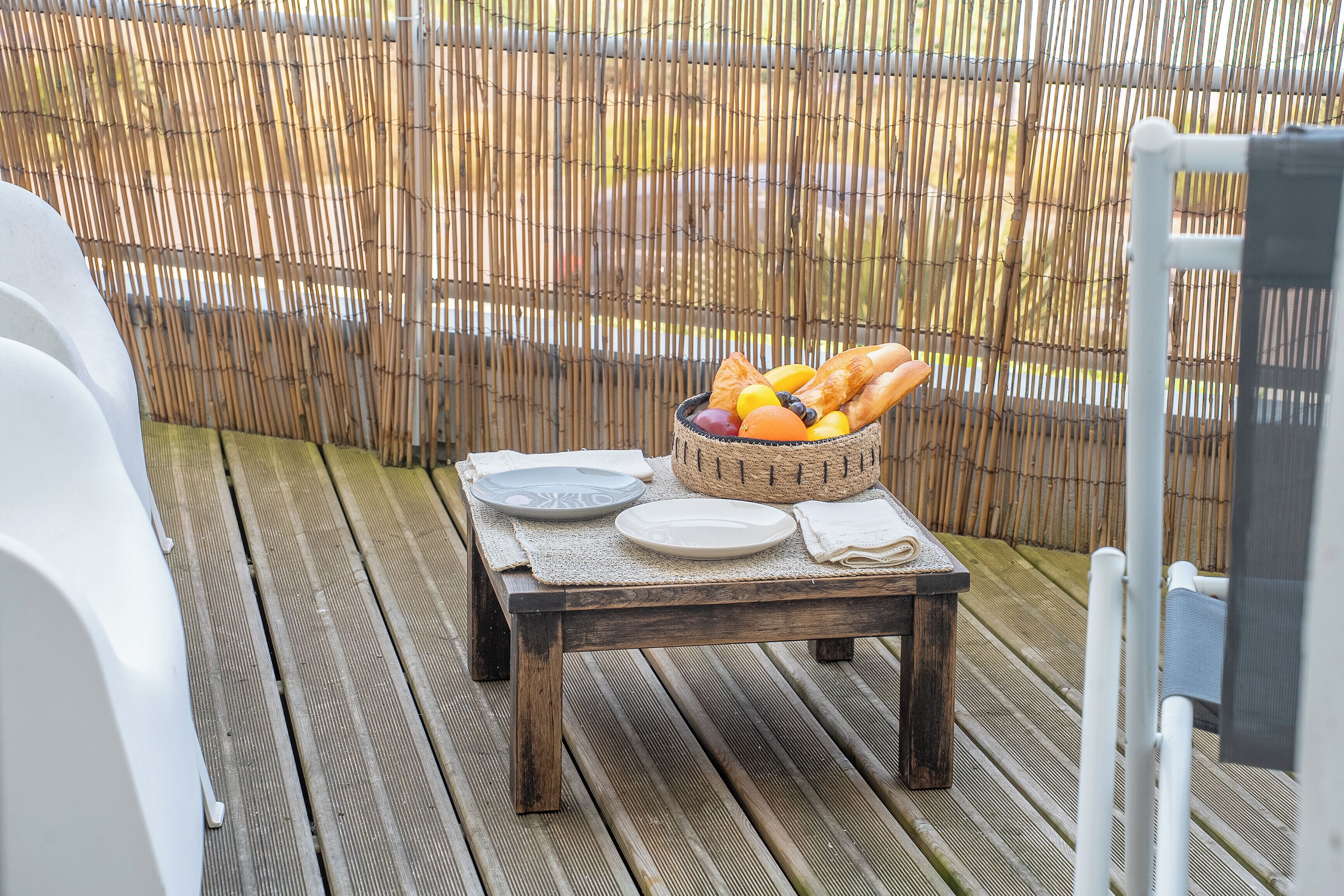 Een houten terras met een eettafel, compleet met borden, een fruitschaal en een geweven mat.
