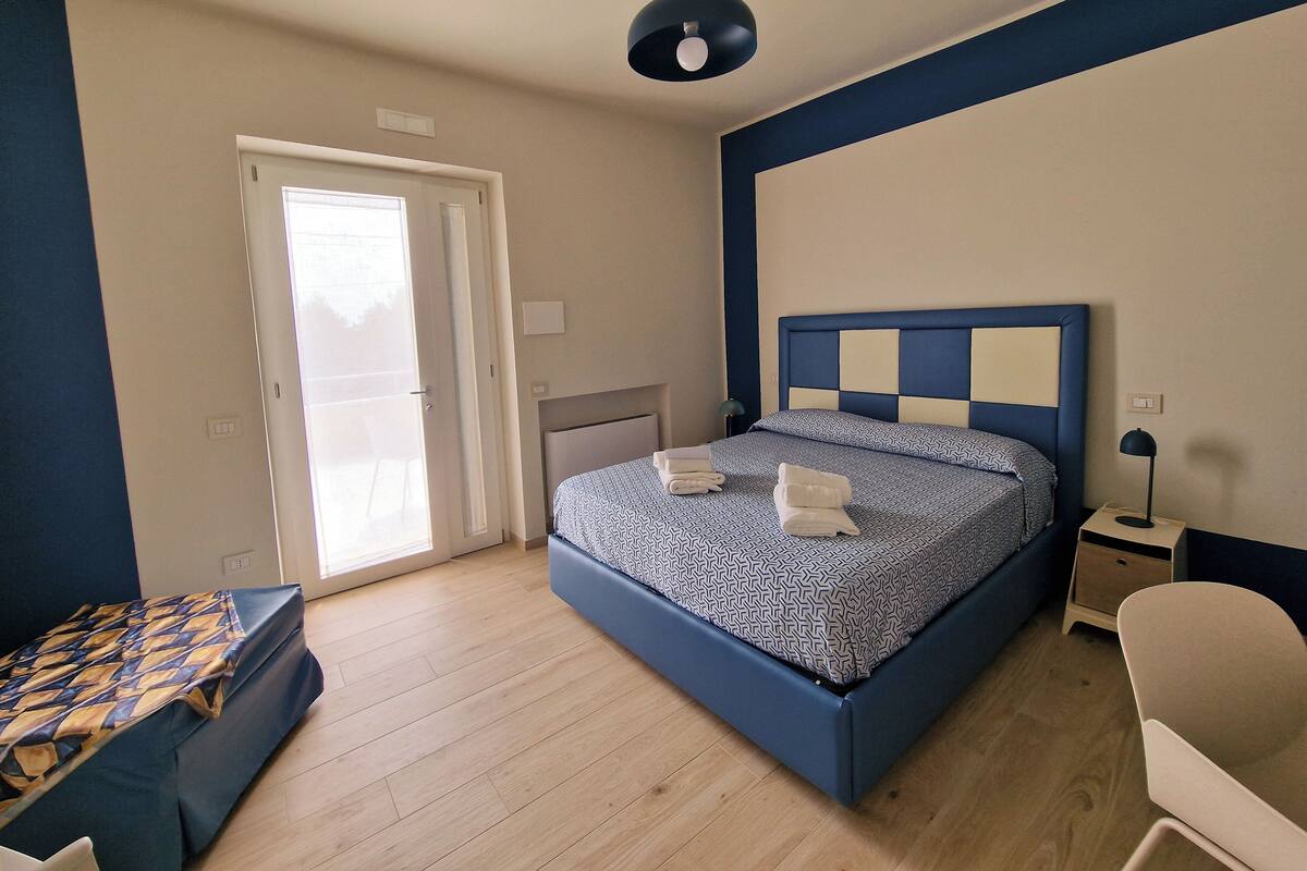 A bedroom with a bed, a desk, a chair, a nightstand, and a door leading to a balcony.
