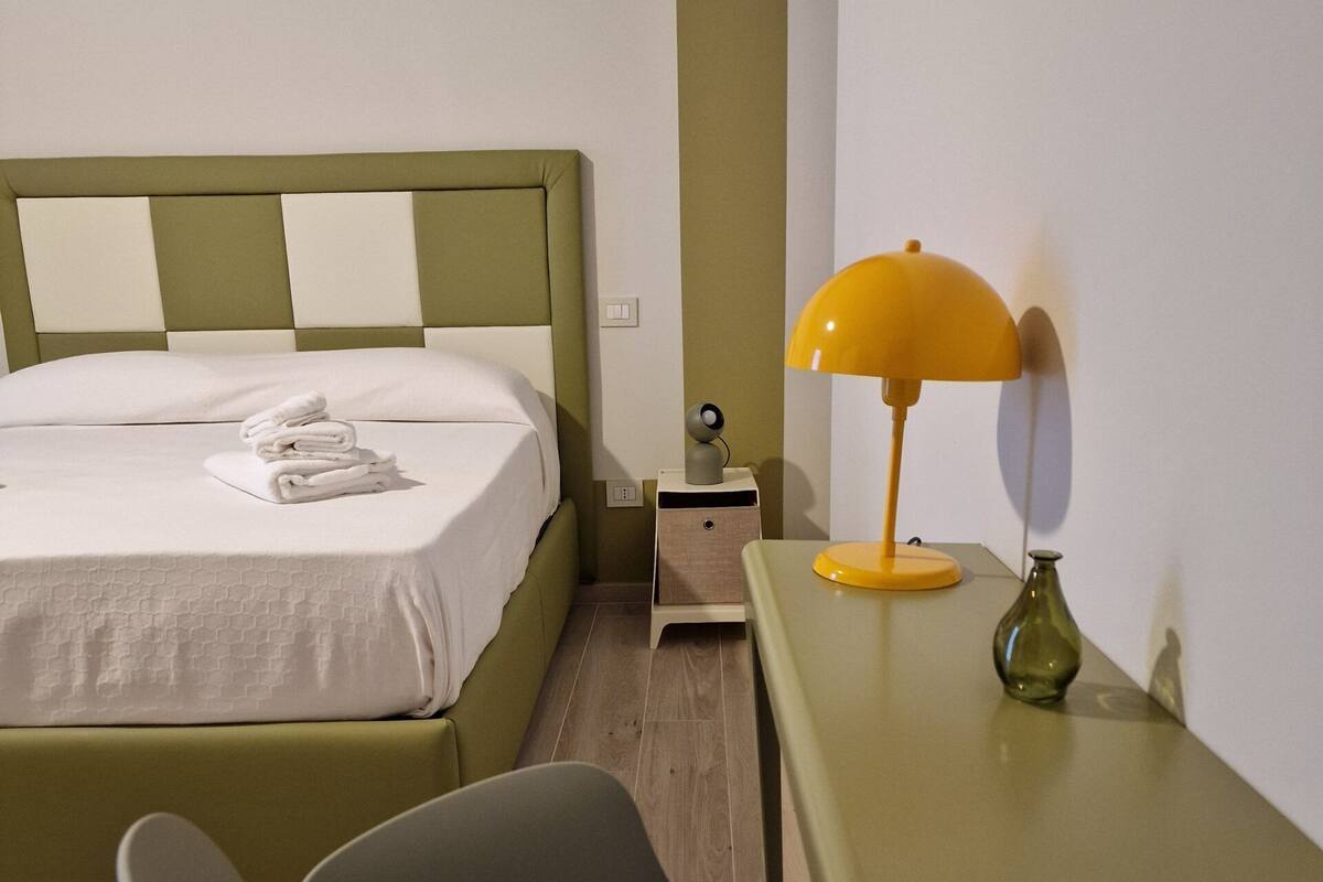 A neatly made bed with a green headboard, a small bedside table with a lamp, and a modern chair.