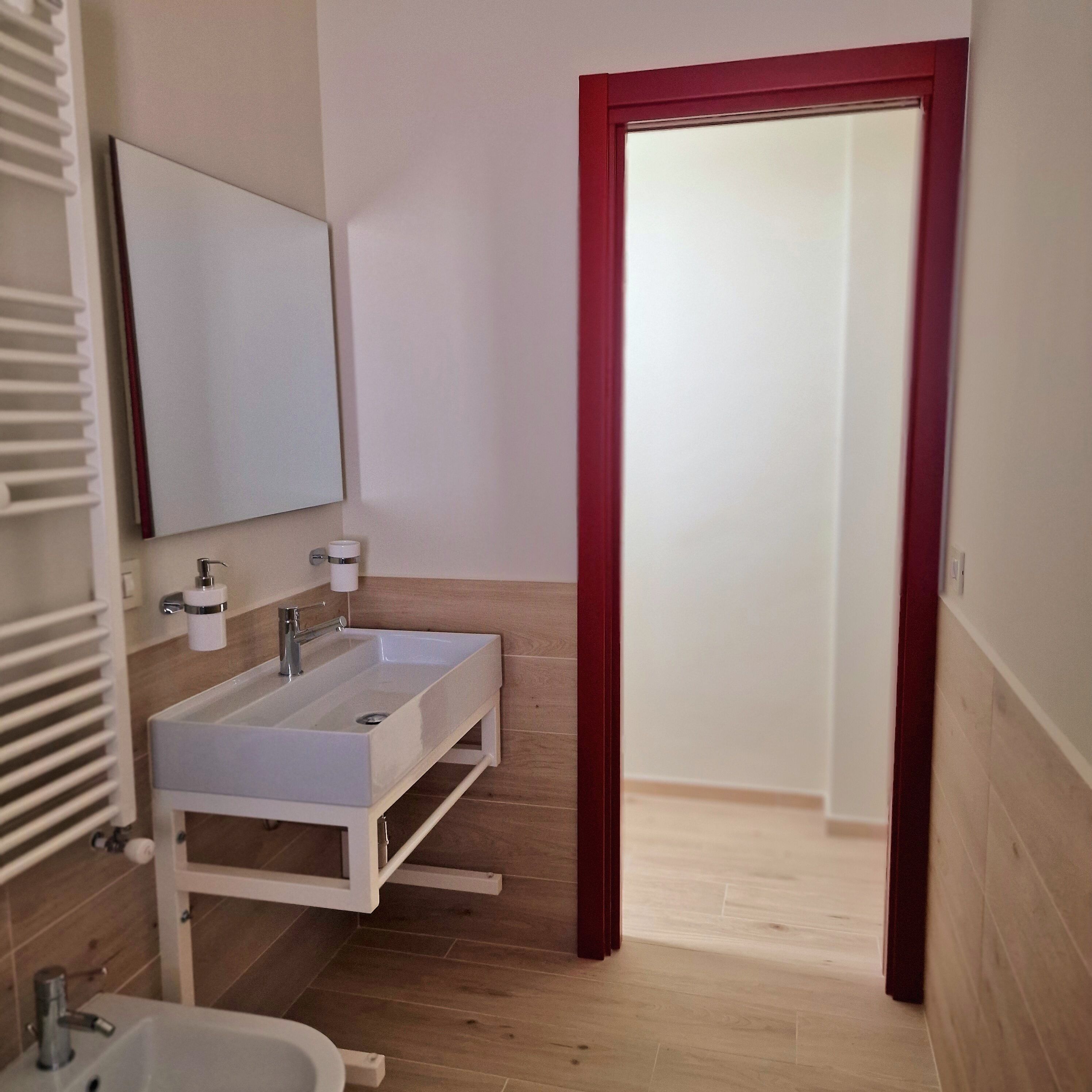 A bathroom with a double sink, a mirror, a toilet, and a red-framed door.
