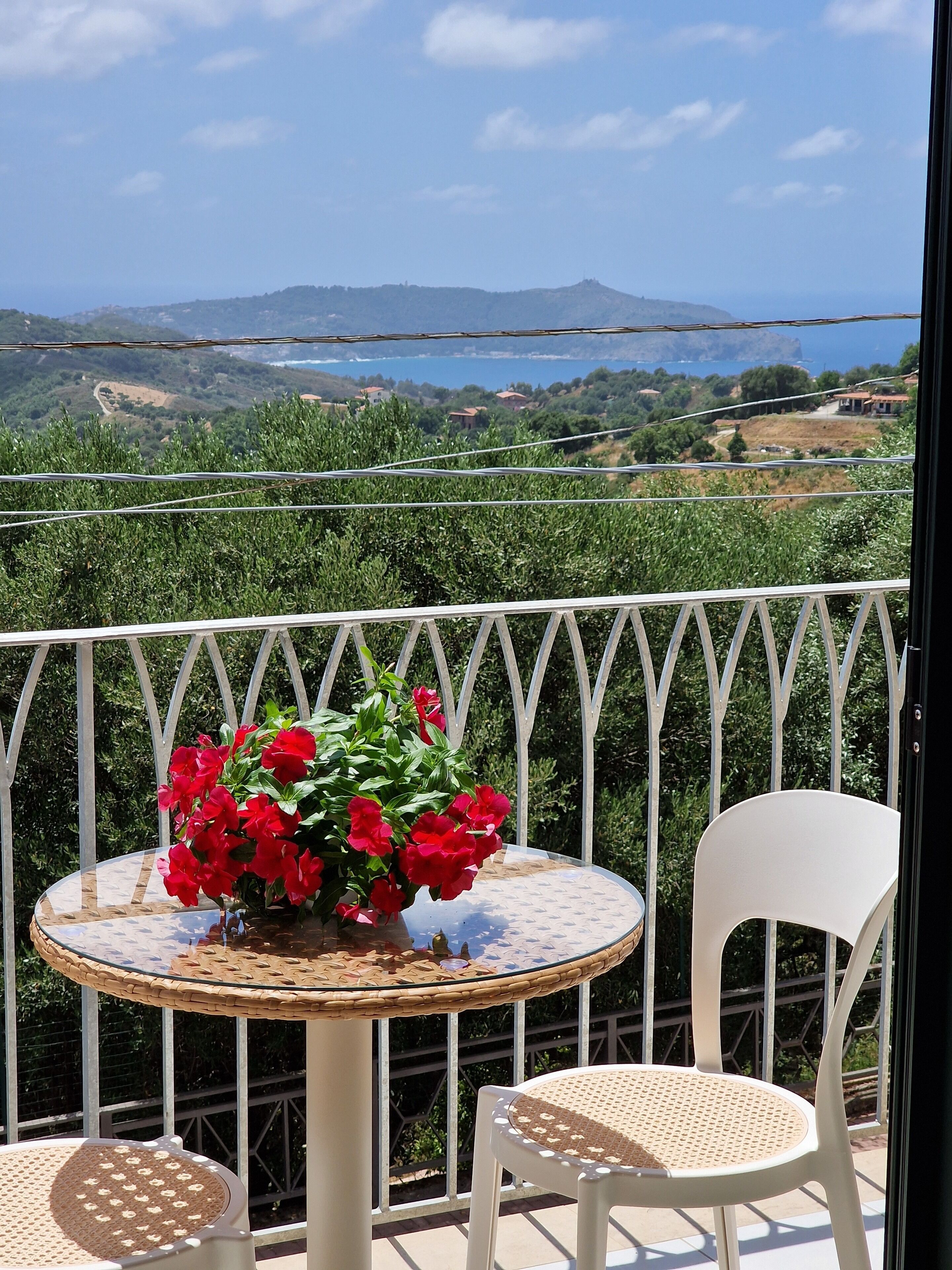 Un balcón con una mesa redonda y dos sillas, un florero con flores rojas, y vista a un paisaje costero.