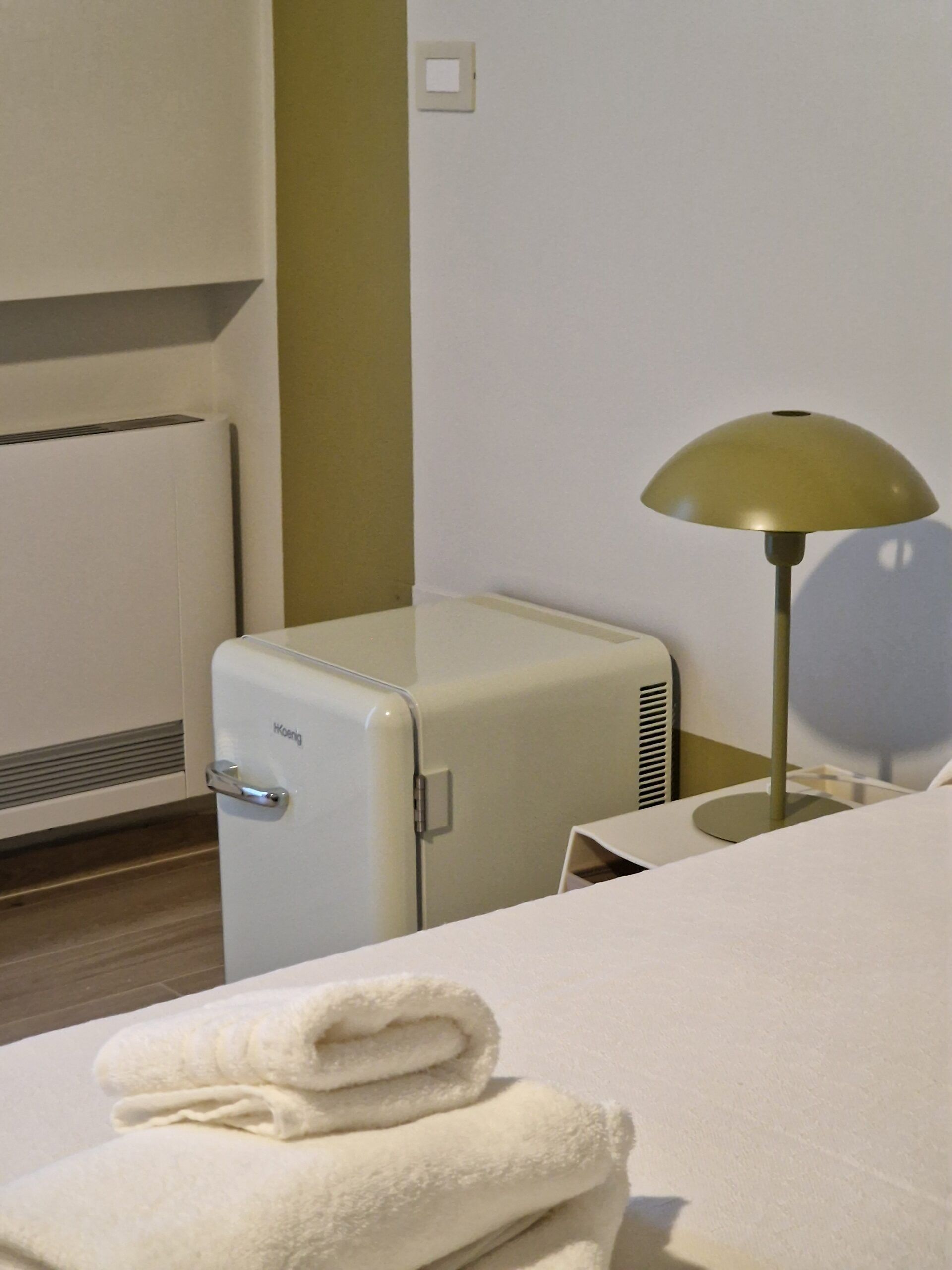 A hotel room with a bed, a small table, a lamp, a heater, and a folded towel.