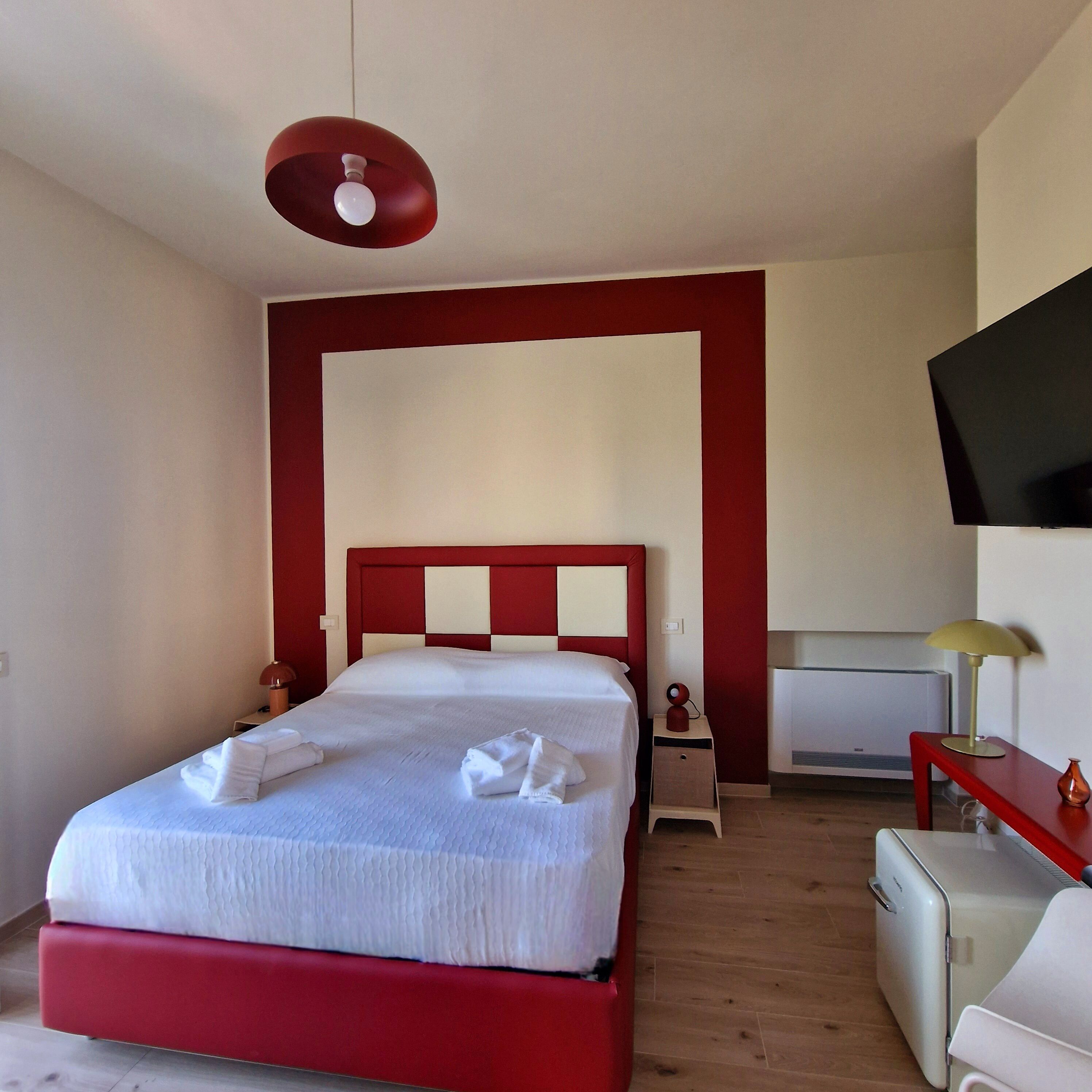 A hotel room with a red and white checkered headboard, a bed with white linens, a red hanging light fixture, a flat-screen TV on the wall, and a small bedside table with a lamp.