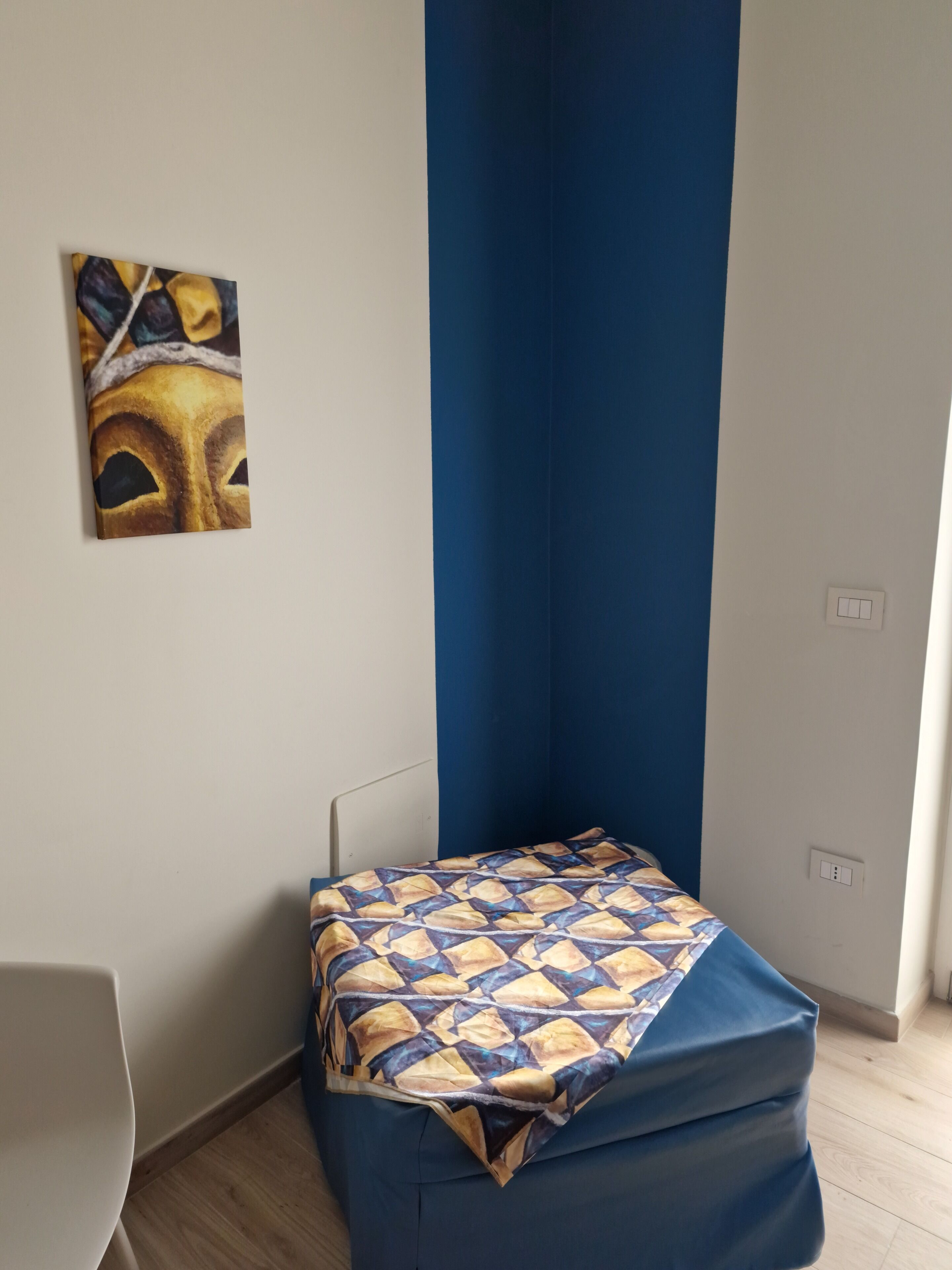 A small room with a bed covered in a patterned blanket, a blue wall, and a painting on the wall.
