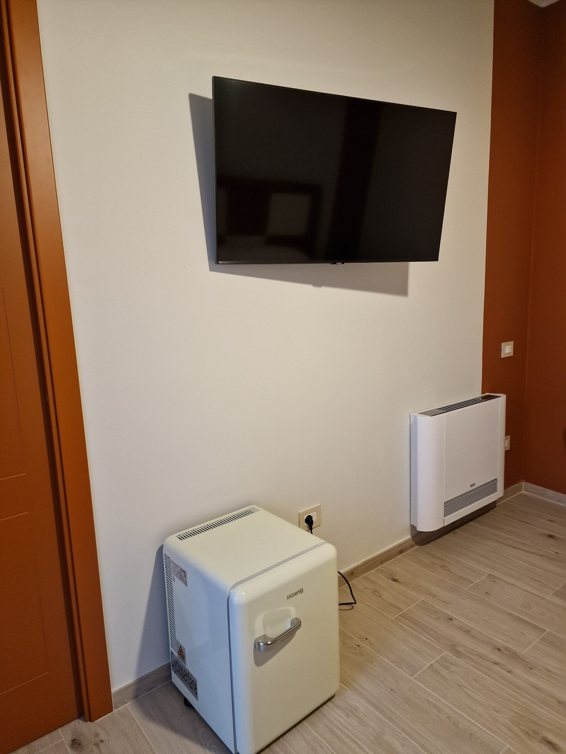 Ett rum med en väggmonterad platt-TV, en vit, portabel luftkonditioneringsapparat och en väggmonterad värmare.
