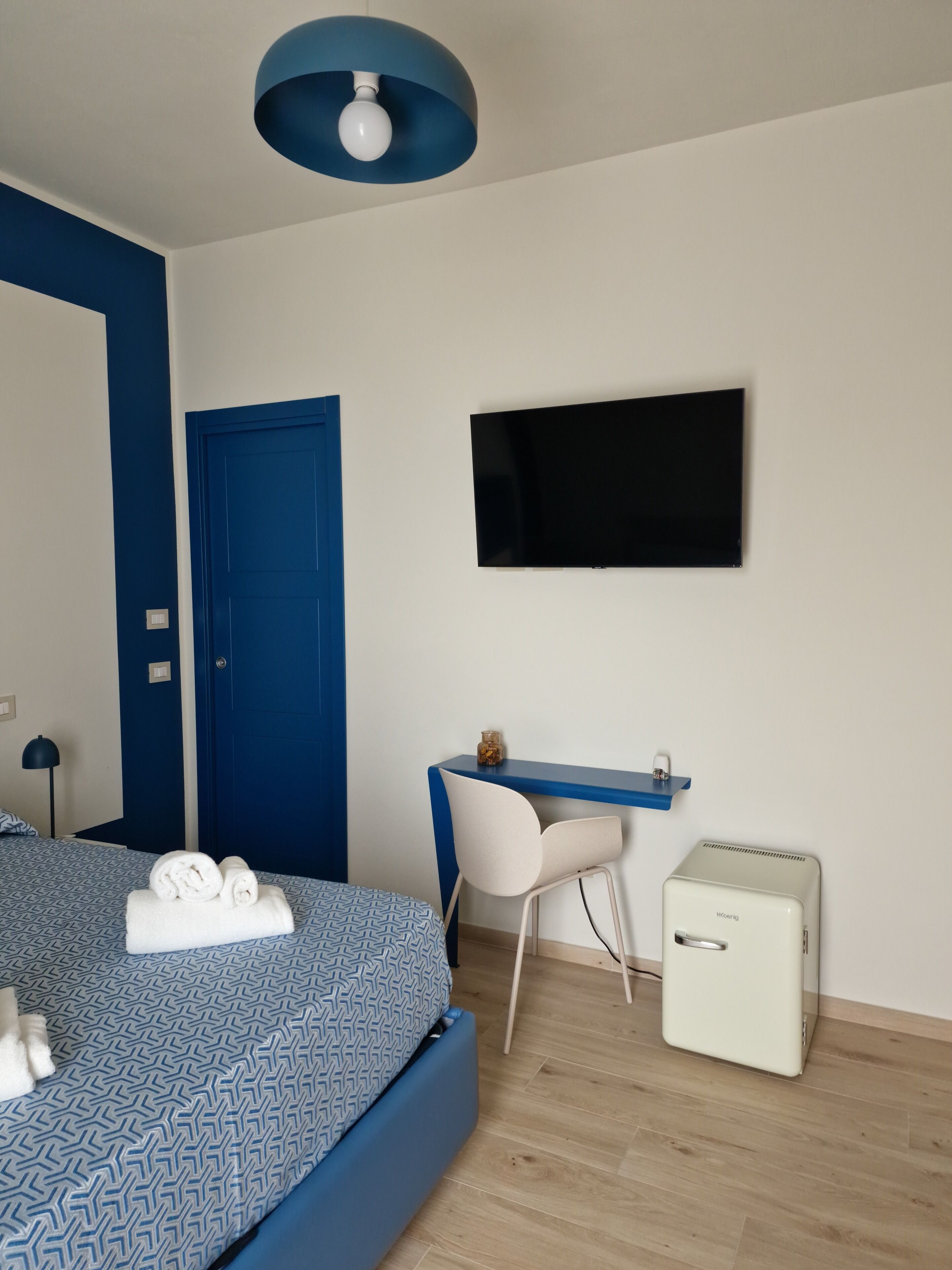 Una habitación de hotel moderna con una decoración azul y blanca, una cama con toallas ordenadas, un escritorio con silla, un televisor y un pequeño armario blanco.