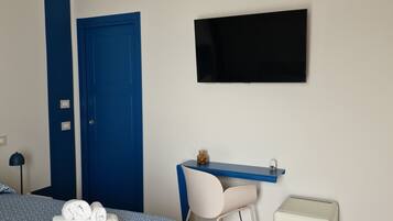 Una habitación de hotel moderna con una decoración azul y blanca, una cama con toallas ordenadas, un escritorio con silla, un televisor y un pequeño armario blanco.