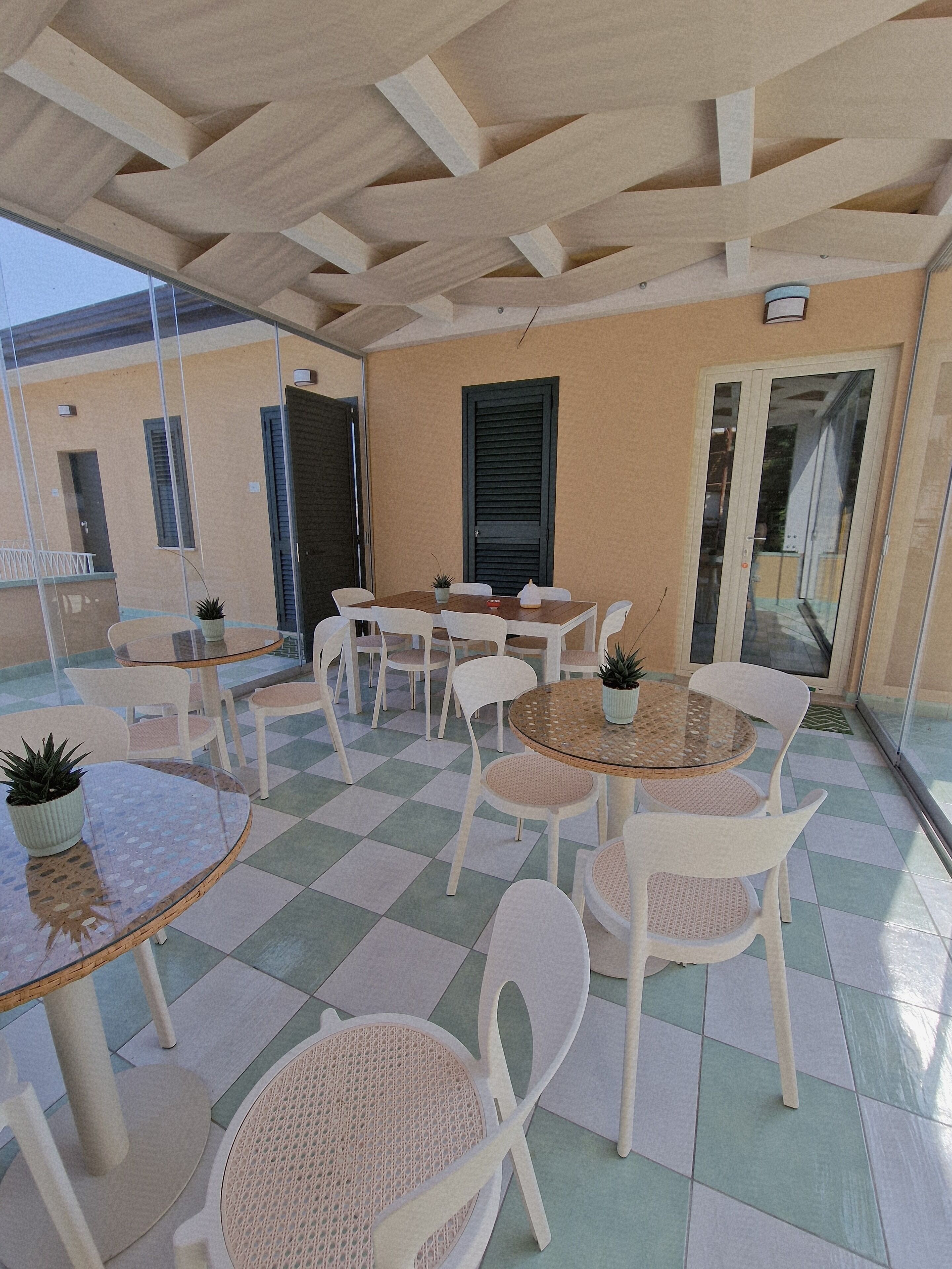 Zona d'estar exterior coberta amb taules i cadires, una planta en test i vista d'un edifici amb porta i finestres.