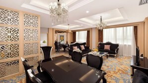 Une chambre d’hôtel comprenant un coin repas, deux canapés, un lustre et une grande fenêtre avec des rideaux.