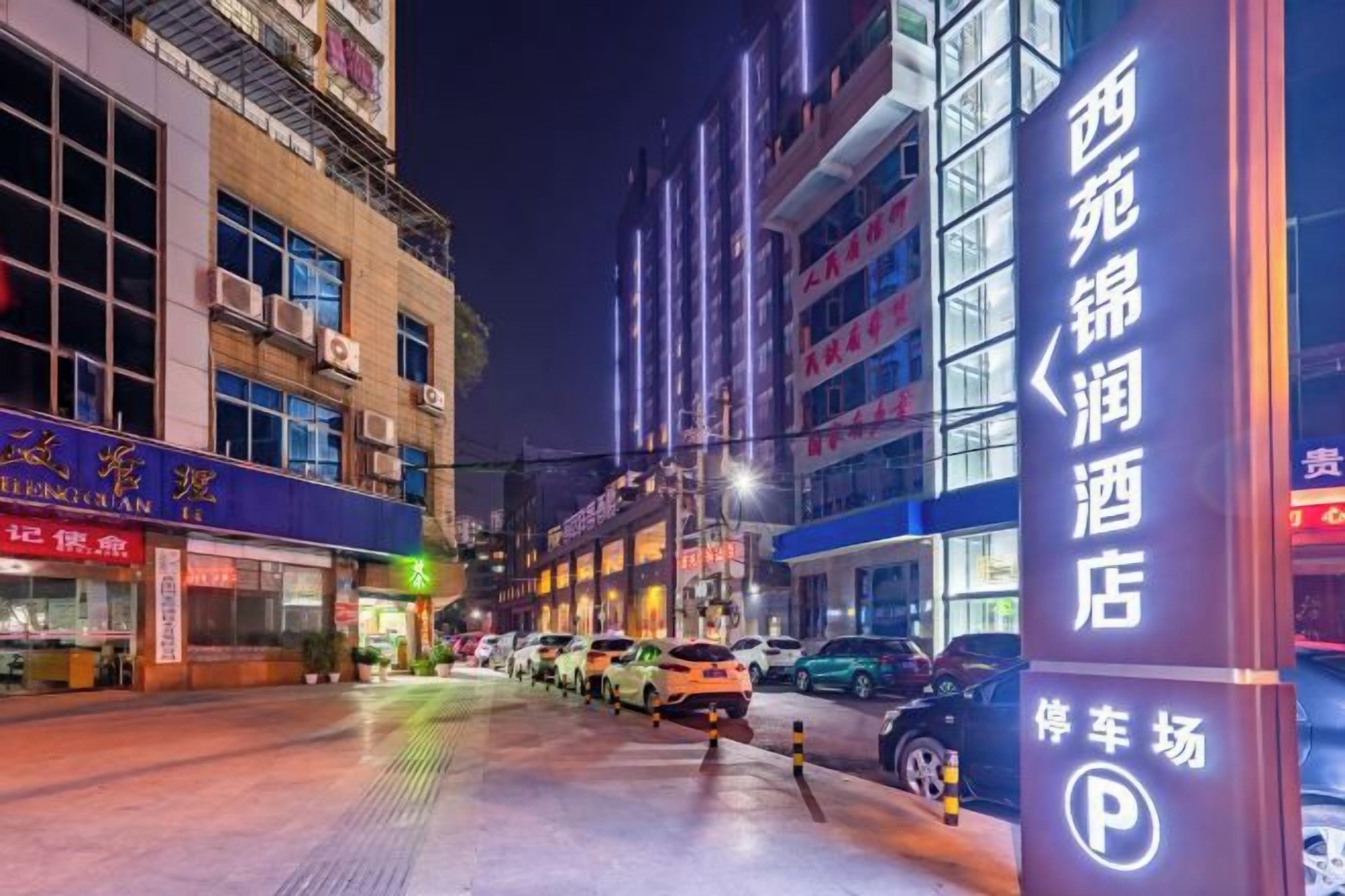 A nighttime street view with a hotel sign, parked cars, and buildings with illuminated signs.