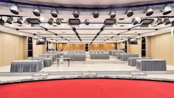 Uma sala de conferência com várias mesas e cadeiras, um palco com tapete vermelho e luminárias no teto.