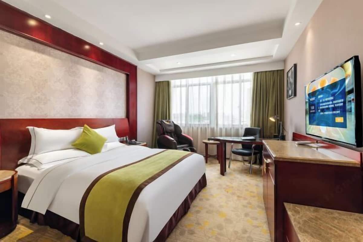 Habitación de hotel con cama, escritorio, silla, televisión y ventanas amplias.