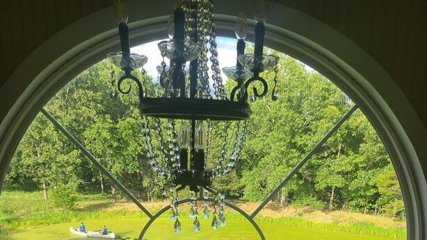 Una stanza con un'ampia finestra ad arco che si affaccia su una zona erbosa con uno stagno e una fila di alberi.