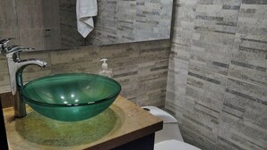 Un baño con un lavamanos de vidrio verde, un inodoro blanco y una toalla colgada en un perchero.