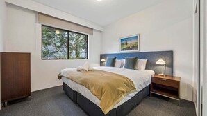 A hotel room with a bed, bedside table, lamp, window, and artwork on the wall.