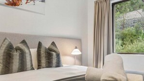 Habitación de hotel con cama, almohadas, lámpara, ventana con cortinas y vista a zonas verdes.