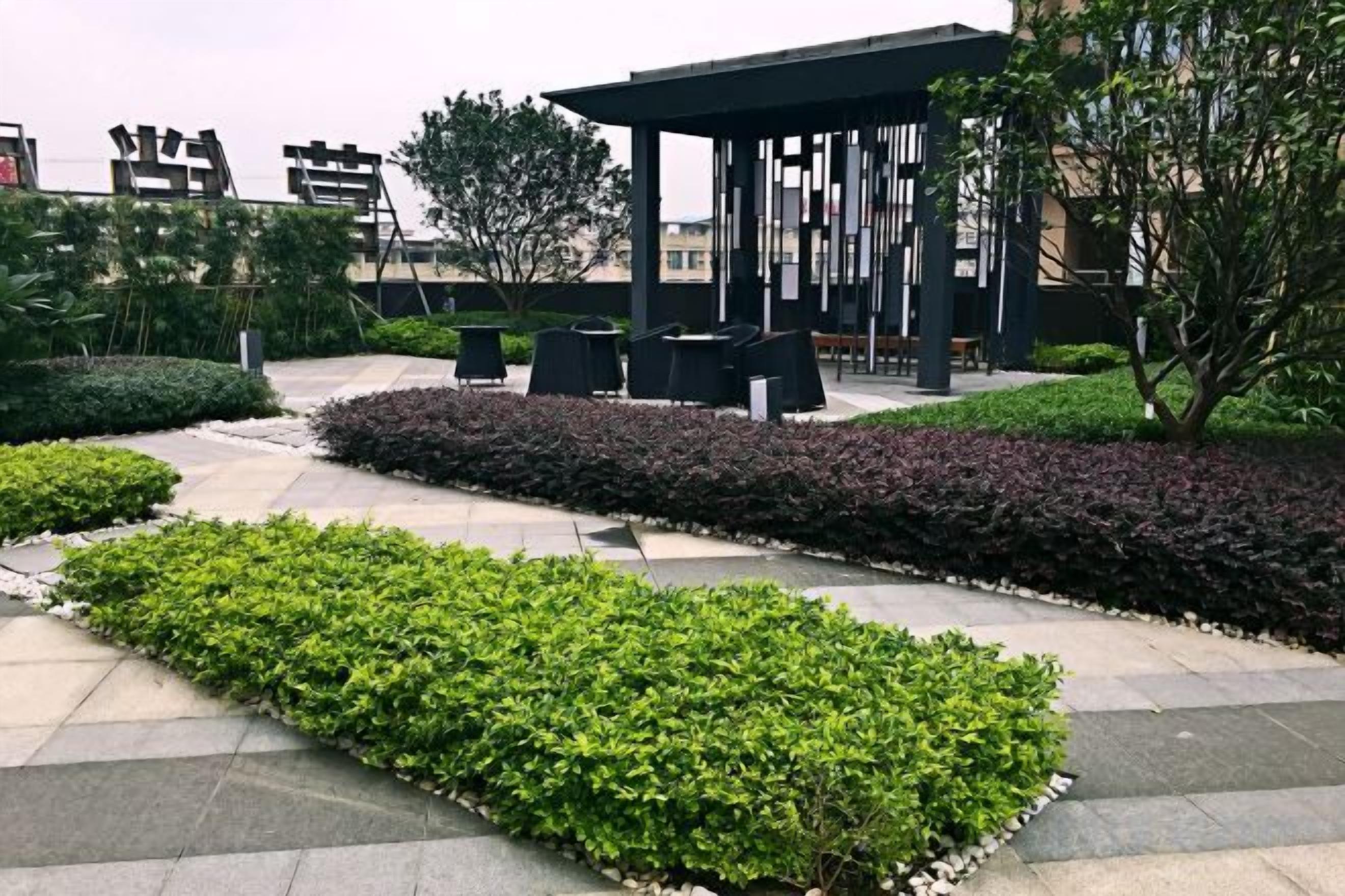 An outdoor seating area with tables and chairs, surrounded by landscaped gardens and a pergola structure.