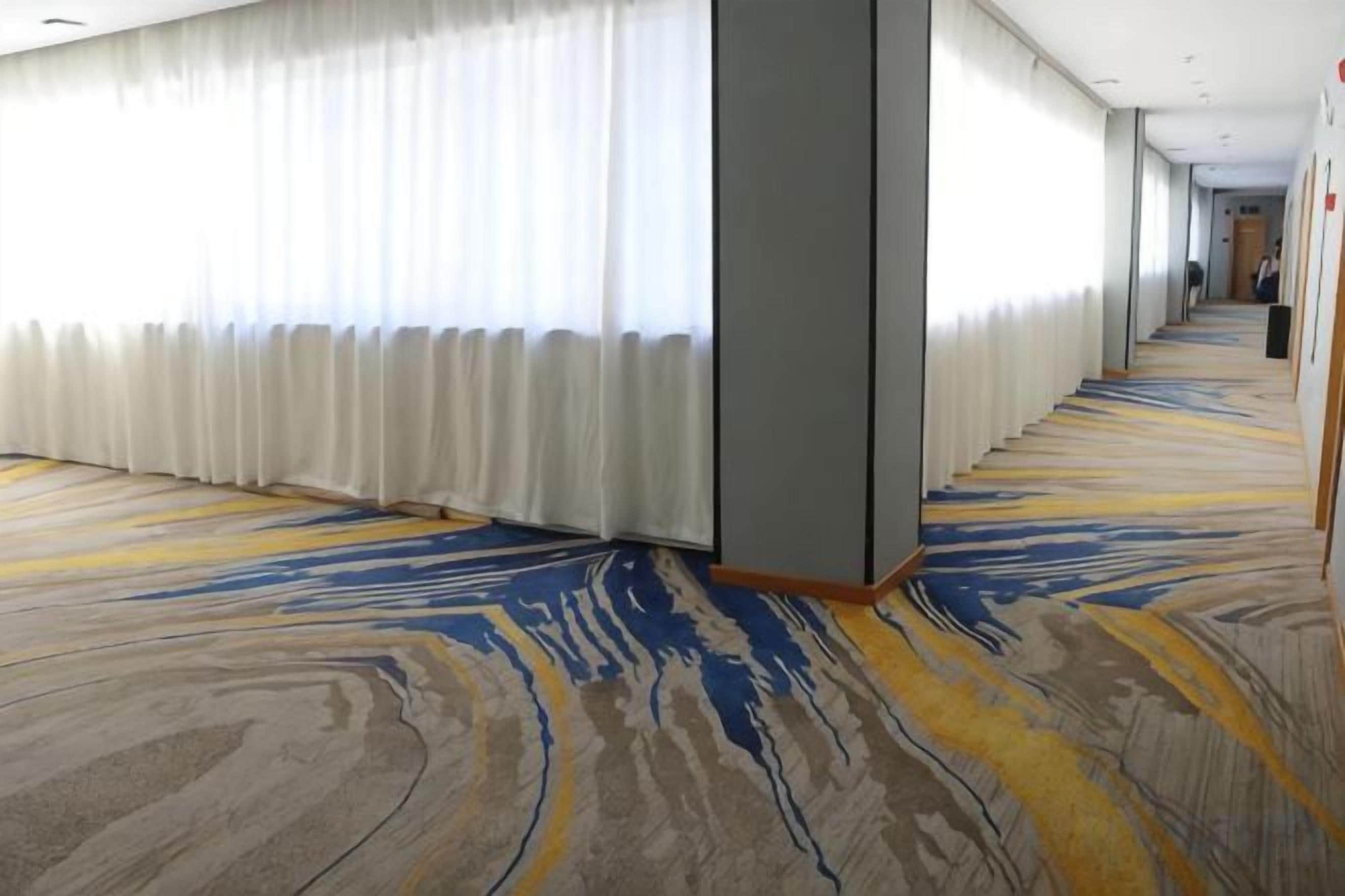 A hotel corridor with a patterned carpet, a large window with white curtains, and a reception desk at the end.