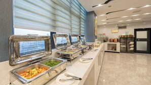 Uma área de buffet com diversas estações de serviço de comida, um balcão com utensílios e uma geladeira ao fundo.