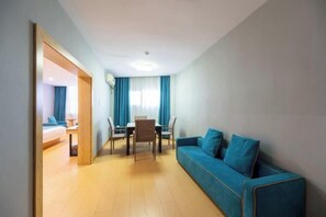 A modern hotel room with a blue sofa, a dining table with chairs, and a separate bedroom area with a bed.